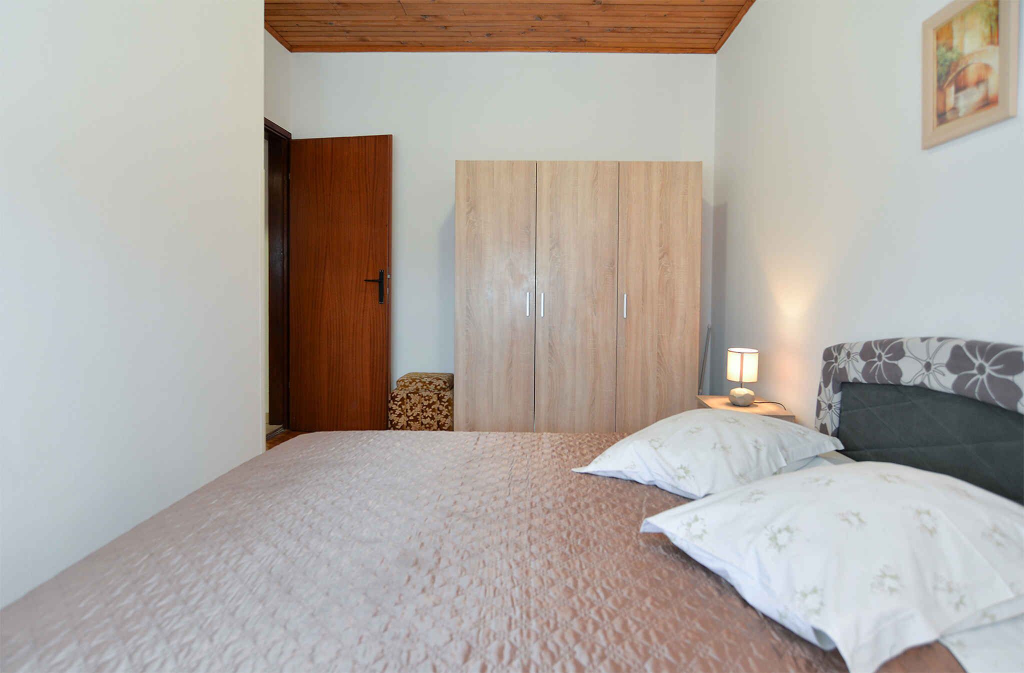 Ein Schlafzimmer mit einem Bett, einem Kleiderschrank, einem Nachttisch mit Lampe und einem Kissen mit Blumenmuster.