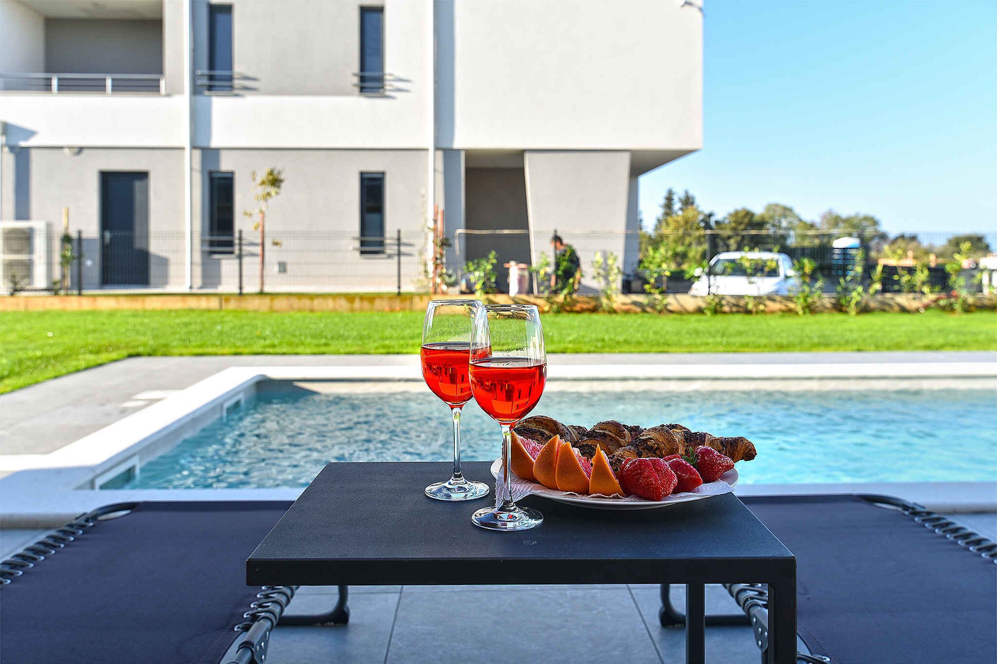 A table with two glasses of red wine and a plate of strawberries and chocolate croissants by a poolside.