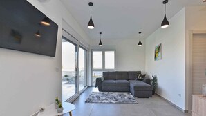 A modern living room with a grey sectional sofa, a small round table with a magazine, and a grey rug. There are pendant lights above the sofa and a large window providing natural light. - Modernly furnished villa with swimming pool (Peroj)