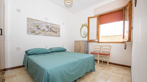 A bedroom with a bed, a chair, a window with a red blind, a mirror, and a wall-mounted artwork.