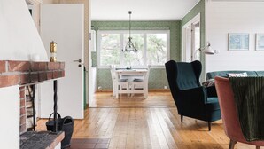 A cozy room with a brick fireplace, wooden flooring, a dining area with a table and chairs, and a green upholstered chair. - Smultronstället by Interhome (Stavsnäs)