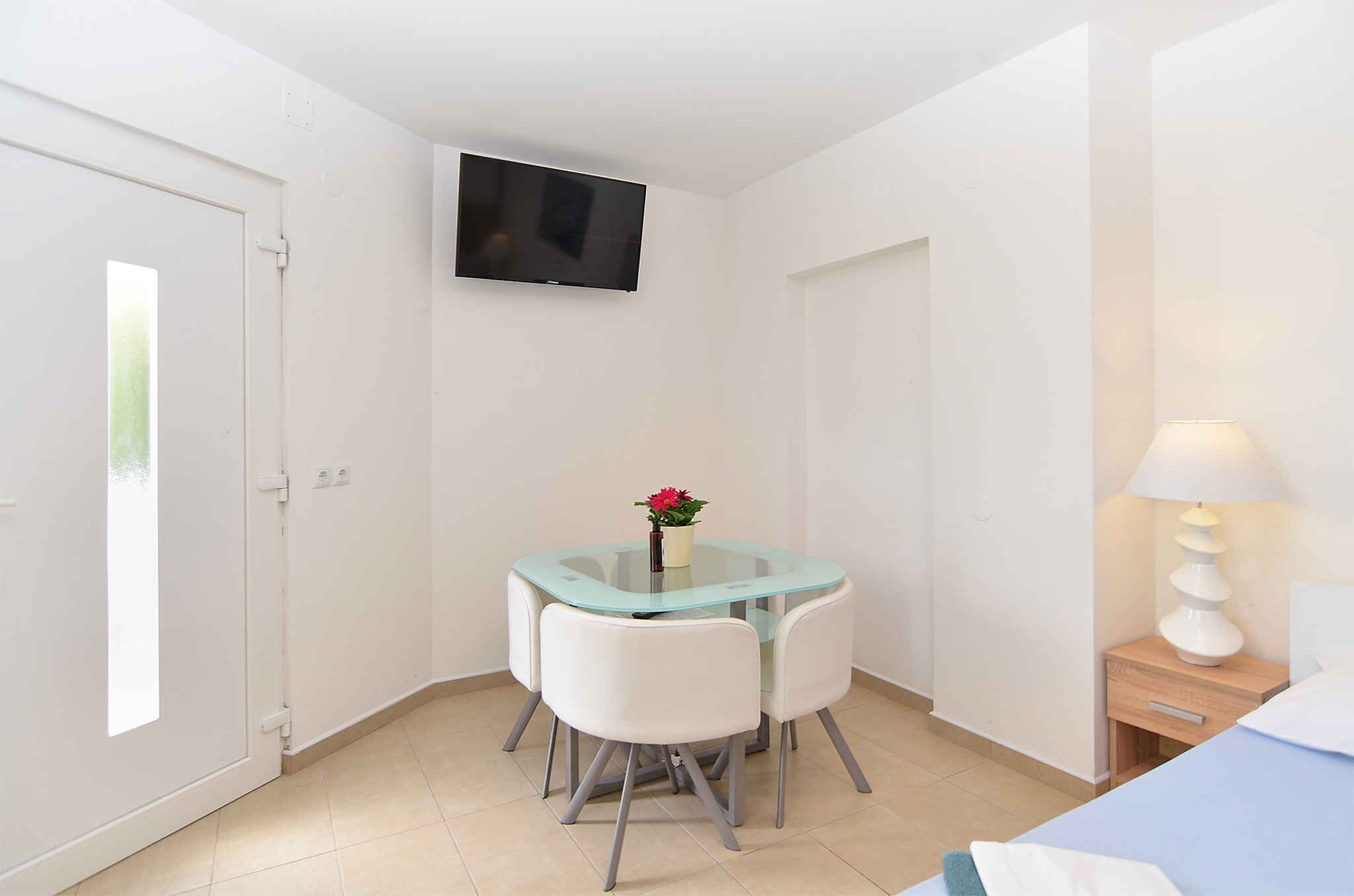 A small, clean room with a glass-top table, white chairs, a TV mounted on the wall, a bedside lamp, and a bed with a blue cover.