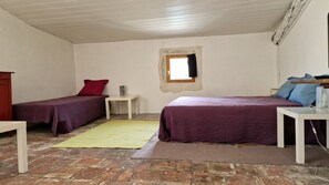 Una habitación con dos camas, una mesita, un ventanal y suelo de piedra.