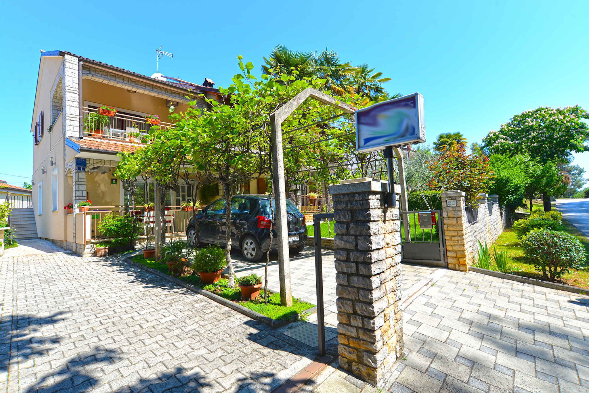 A residential building with a paved driveway, a car parked in front, and a garden with various plants and flowers.