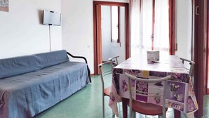 A room with a blue sofa, a table with a floral tablecloth, and a TV mounted on the wall. - Holiday apartment with balcony and air conditioning (Lido di Jesolo)