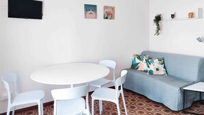 A room with a round white table and four white chairs, a light blue sofa with a patterned cushion, and a wall-mounted TV. - Studio only 100 m from the beach (Lido di Jesolo)