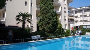 A swimming pool with a white fence, a palm tree, and a tall evergreen tree. - Studio only 100 m from the beach (Lido di Jesolo)