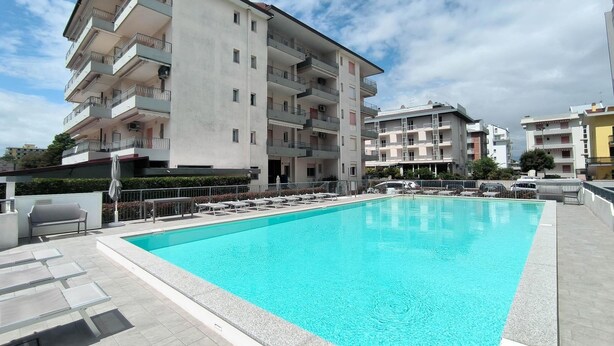 A swimming pool with lounge chairs and a building with multiple balconies in the background. - Studio only 100 m from the beach (Lido di Jesolo)