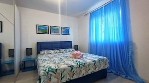 A bedroom with a bed, a blue headboard, a window with blue curtains, and two framed pictures on the wall.
