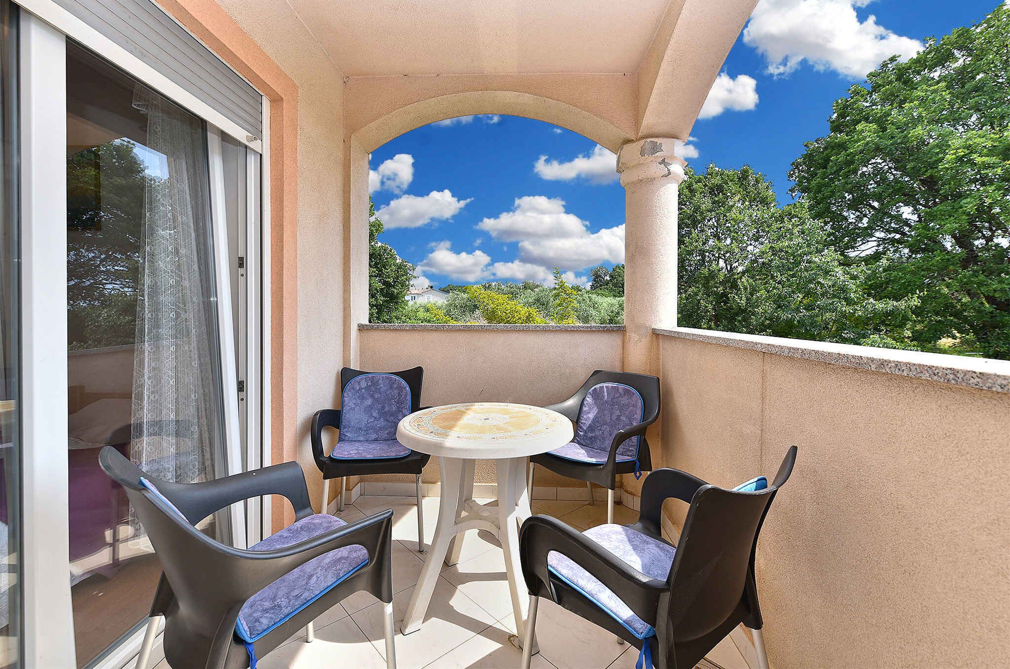A balcony with a table and four chairs, a glass door, and a view of trees and sky.