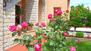Un bâtiment en pierre avec une entrée surmontée de tuiles rouges et un jardin où poussent des rosiers roses.