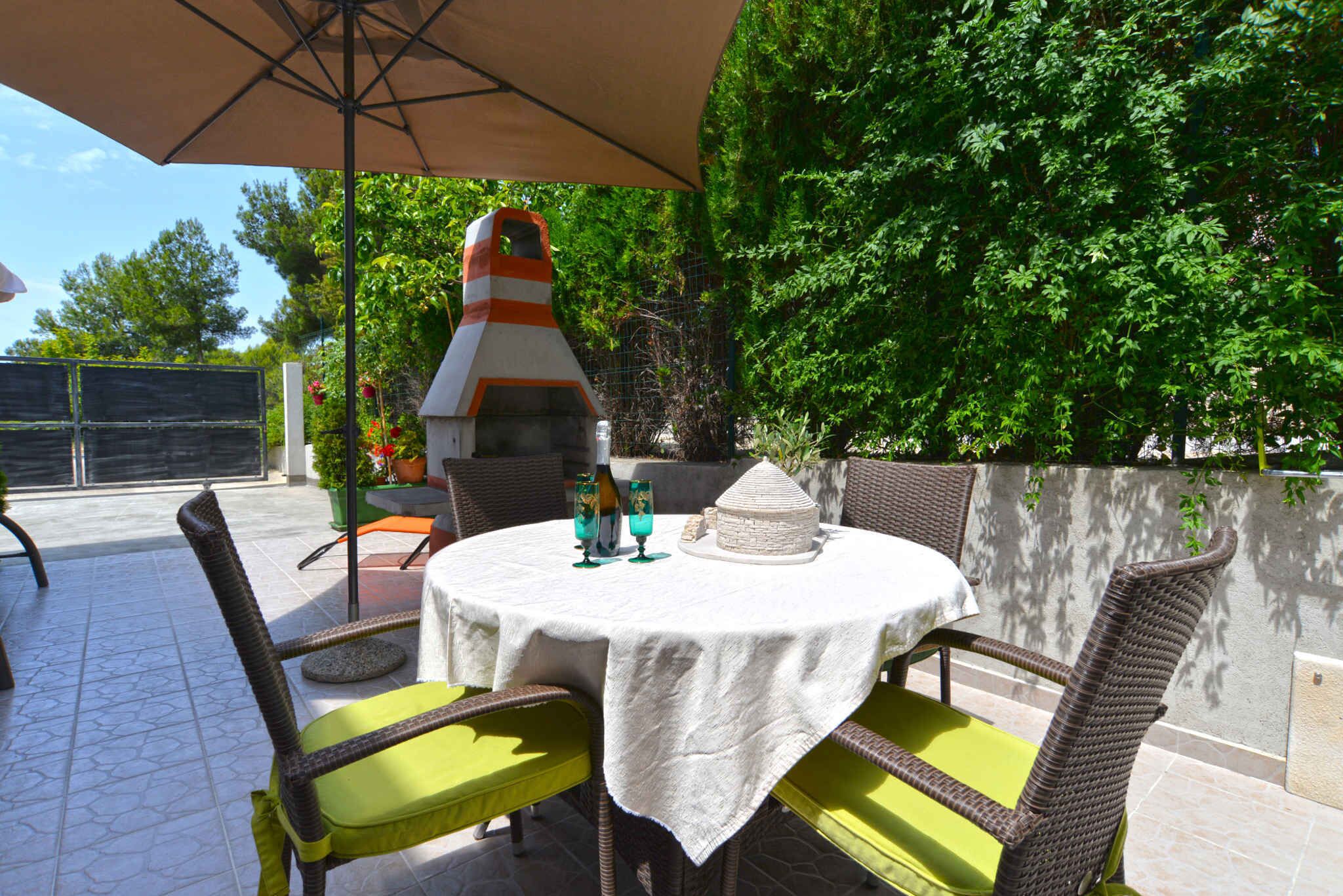 Une terrasse extérieure avec une table ronde recouverte d’une nappe blanche, entourée de chaises en osier avec des coussins verts.