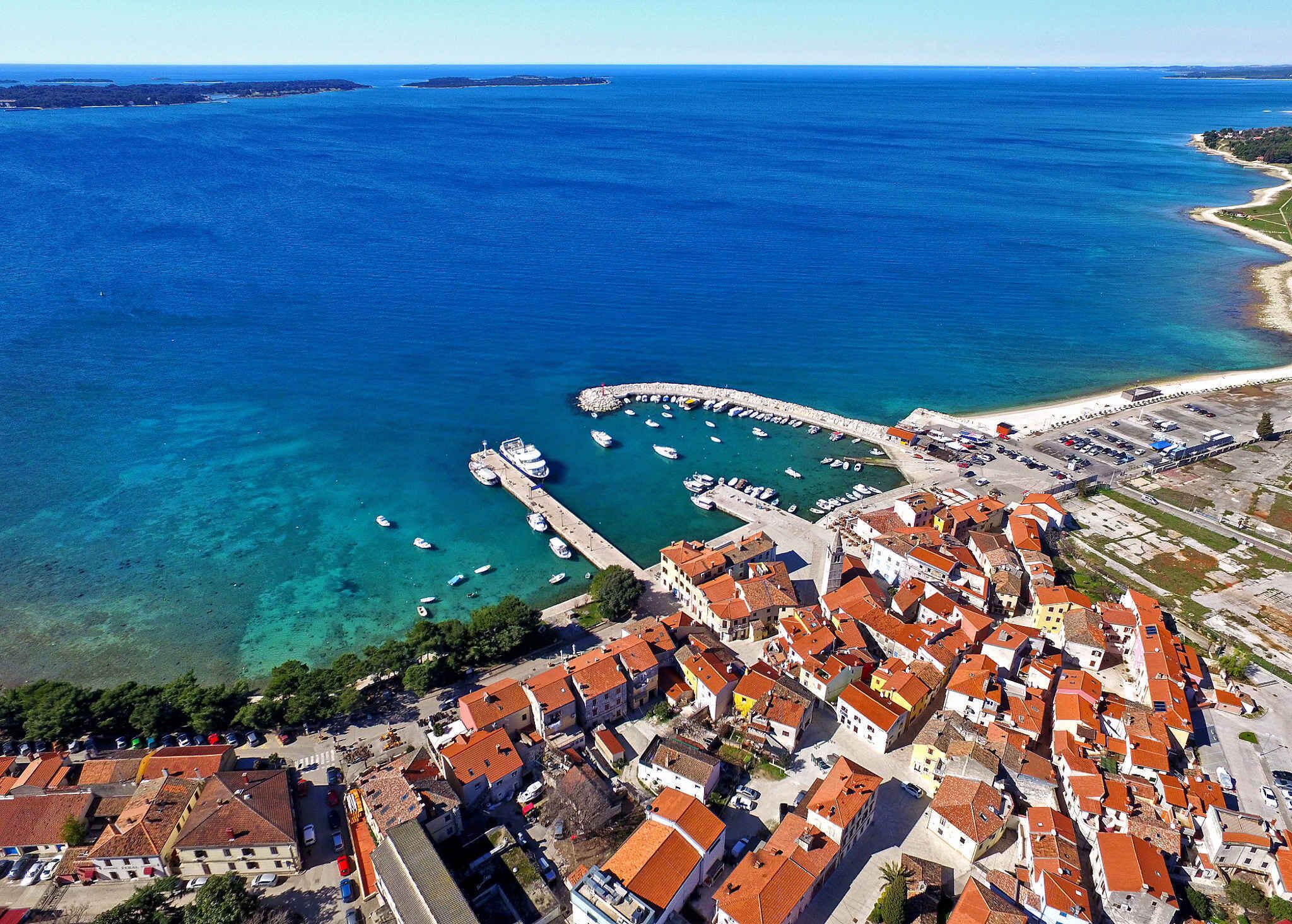 An aerial view of a coastal town with a marina, clear blue waters, and traditional red-tiled buildings.