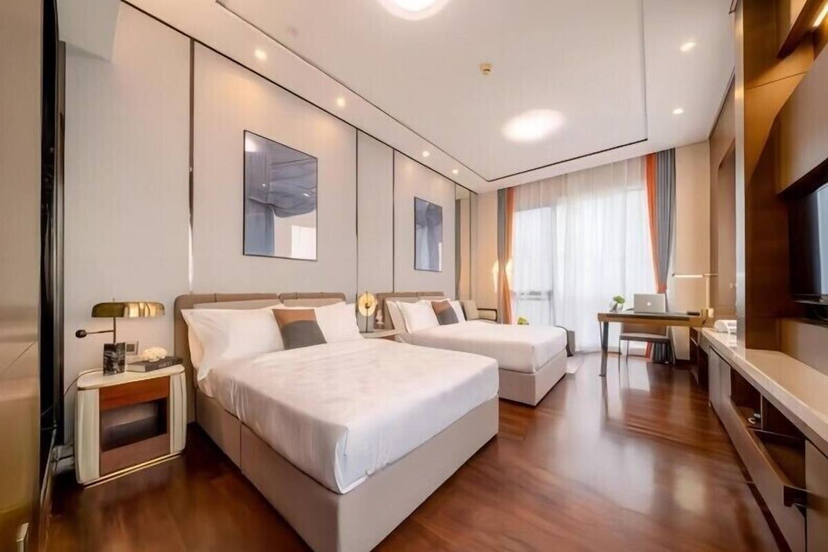 A hotel room with two beds, a desk, a TV, and a large window with curtains.