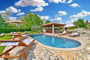 A swimming pool with lounge chairs and umbrellas, surrounded by stone tiles and greenery. - Villa with swimming pool, playground and garden kitchen (Tinjan)