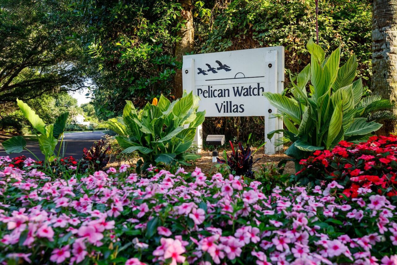 Une enseigne indiquant l’emplacement de « Pelican Watch Villas », entourée d’une végétation luxuriante et de fleurs roses.