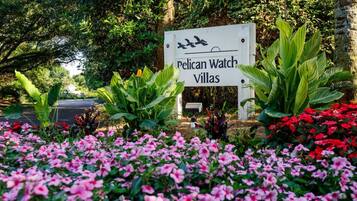 Une enseigne indiquant l’emplacement de « Pelican Watch Villas », entourée d’une végétation luxuriante et de fleurs roses.