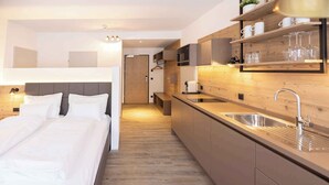 A modern hotel room with a bed, a kitchenette, and a wooden floor.