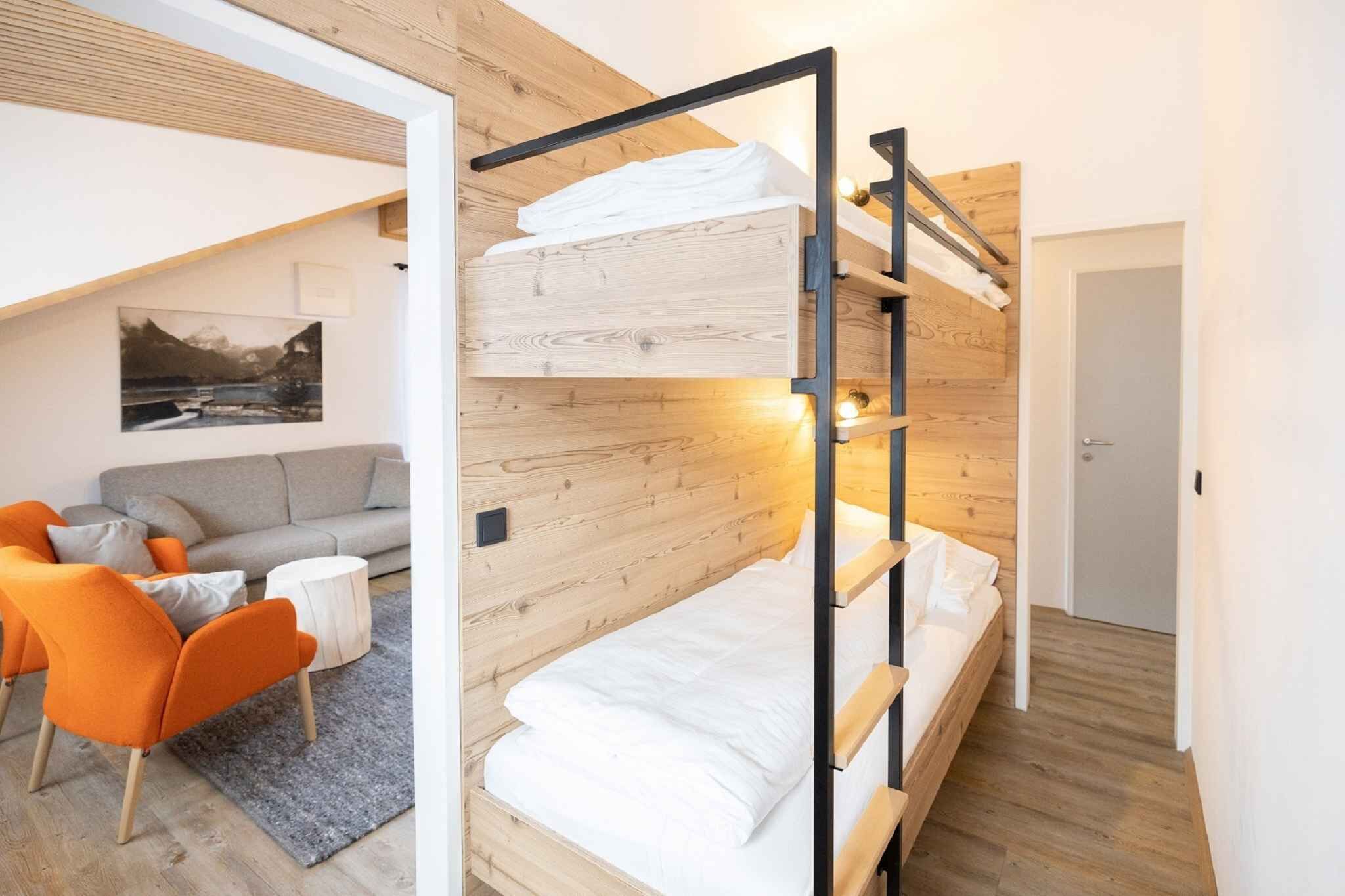 A bunk bed with a wooden headboard, a grey sofa, orange chairs, and a small round table in a room with wooden walls and a picture on the wall.