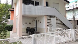 Un bâtiment à deux étages doté d’un balcon à l’étage supérieur, d’un escalier menant à l’étage, et d’un petit espace extérieur aménagé avec des tables et des chaises.