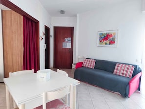 A small, well-lit room with a white table, a grey sofa with red cushions, a wooden door, and a framed painting on the wall. - Holiday apartment with balcony and air conditioning (Lido di Jesolo)