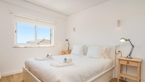 Ein schlicht eingerichtetes Schlafzimmer mit einem großen Bett, zwei Nachttischen, einem Beistelltisch und einem Fenster mit Blick auf Gebäude.