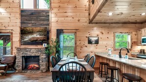 Interior de una cabaña de madera con chimenea de piedra, mesa de comedor, sillas, taburetes de bar y zona de cocina.
