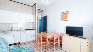 A compact room with a kitchenette, a dining table with chairs, a television, and a patterned floor. - Holiday apartment near the sea (Lido di Jesolo)