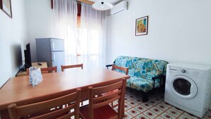A compact living space with a dining table, chairs, a sofa, a washing machine, and a television. - Holiday apartment near the sea (Lido di Jesolo)