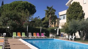 Piscina com espreguiçadeiras e guarda-sóis, cercada por árvores e um edifício.