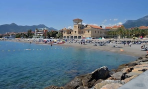 A coastal town with a prominent building featuring a tower, a sandy beach, and rocky shoreline.
