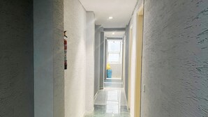 Ein schmaler, gut beleuchteter Flur mit einem Feuerlöscher links, einem Fenster am Ende und einer blauen Mülltonne in der Ferne.