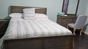 Een slaapkamer met een houten bed, nachtkastje, dressoir, stoel en spiegel.