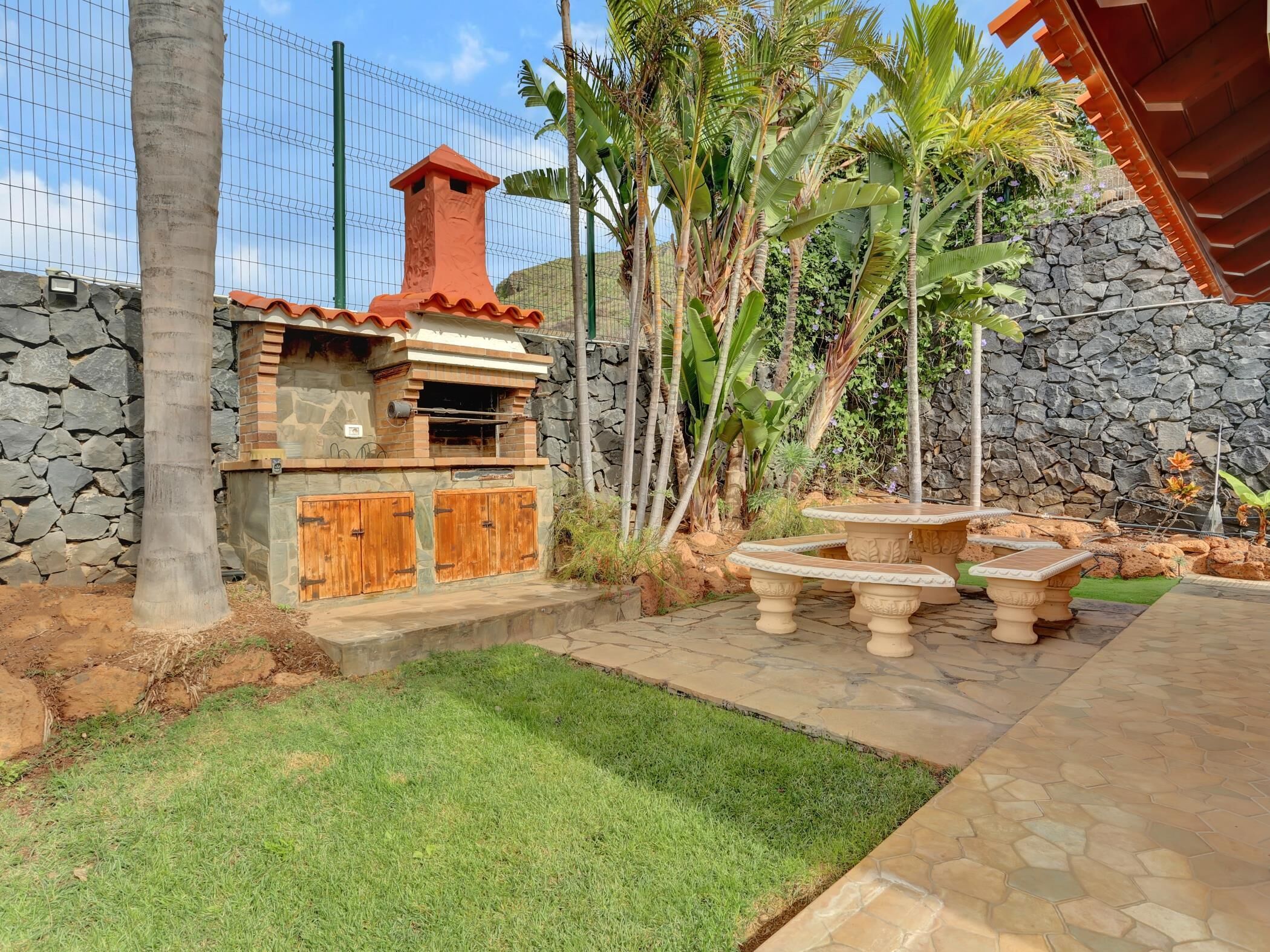 Un patio trasero con un muro de piedra, una zona de barbacoa de ladrillo y un espacio de estar con mesas de piedra.