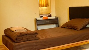 A single bed with a brown mattress and pillows, a bedside table with a lamp, and a mirror above the table. - The House Hostel (Bangkok)