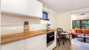 Una cocina moderna con armarios blancos, encimera de madera y lavavajillas integrado. Hay una zona de comedor con mesa y sillas, y vistas al exterior a través de amplios ventanales.