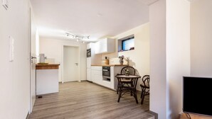 Una cocina moderna con armarios blancos, un comedor con mesa y sillas, un mueble para TV y una ventana con persianas.
