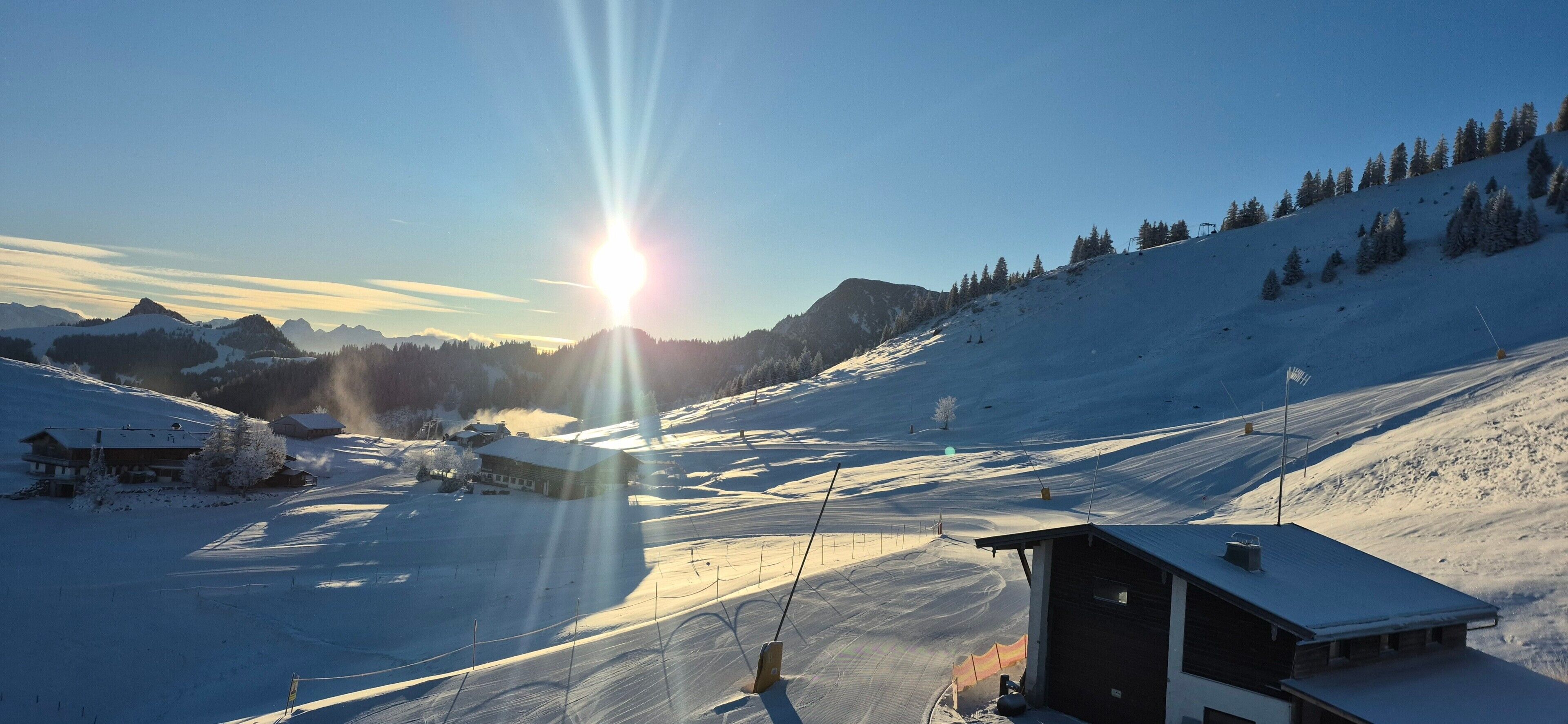 A snow-covered ski resort with a clear sky, a sunlit mountain, and a few buildings.