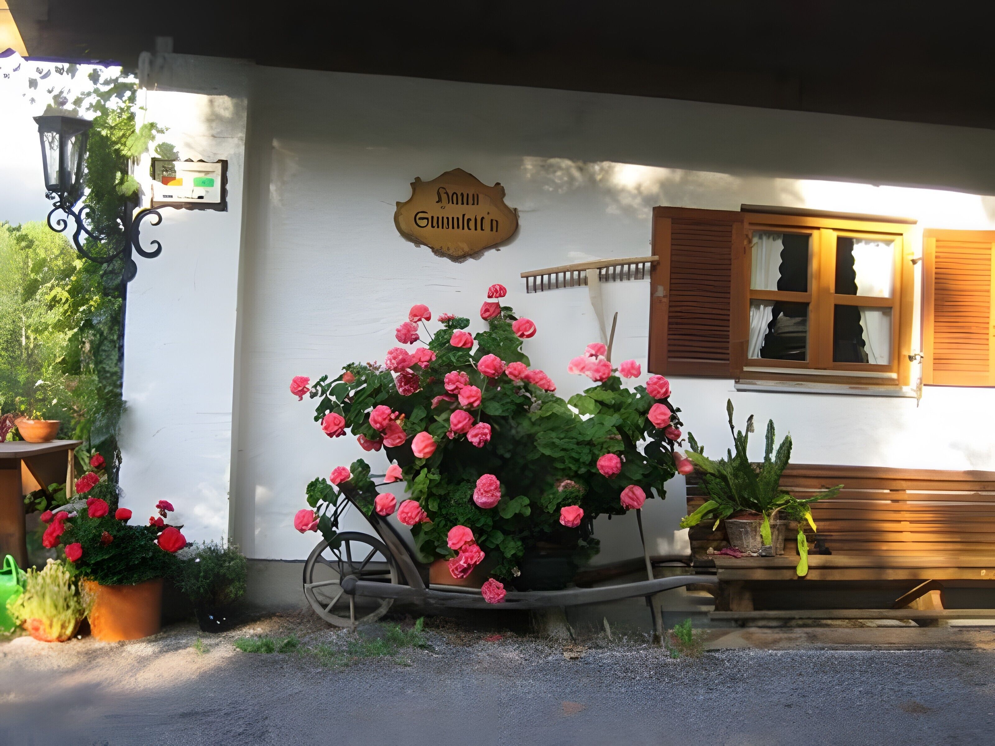 A white building with a sign reading 'Haus Gummeltin' and a wheelbarrow full of pink flowers.