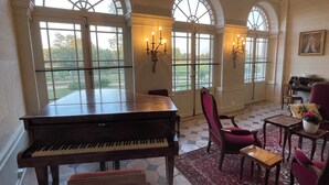 Un piano à queue dans une pièce spacieuse, dotée de grandes fenêtres en arc de cercle, avec un fauteuil rouge, une petite table et un tapis.