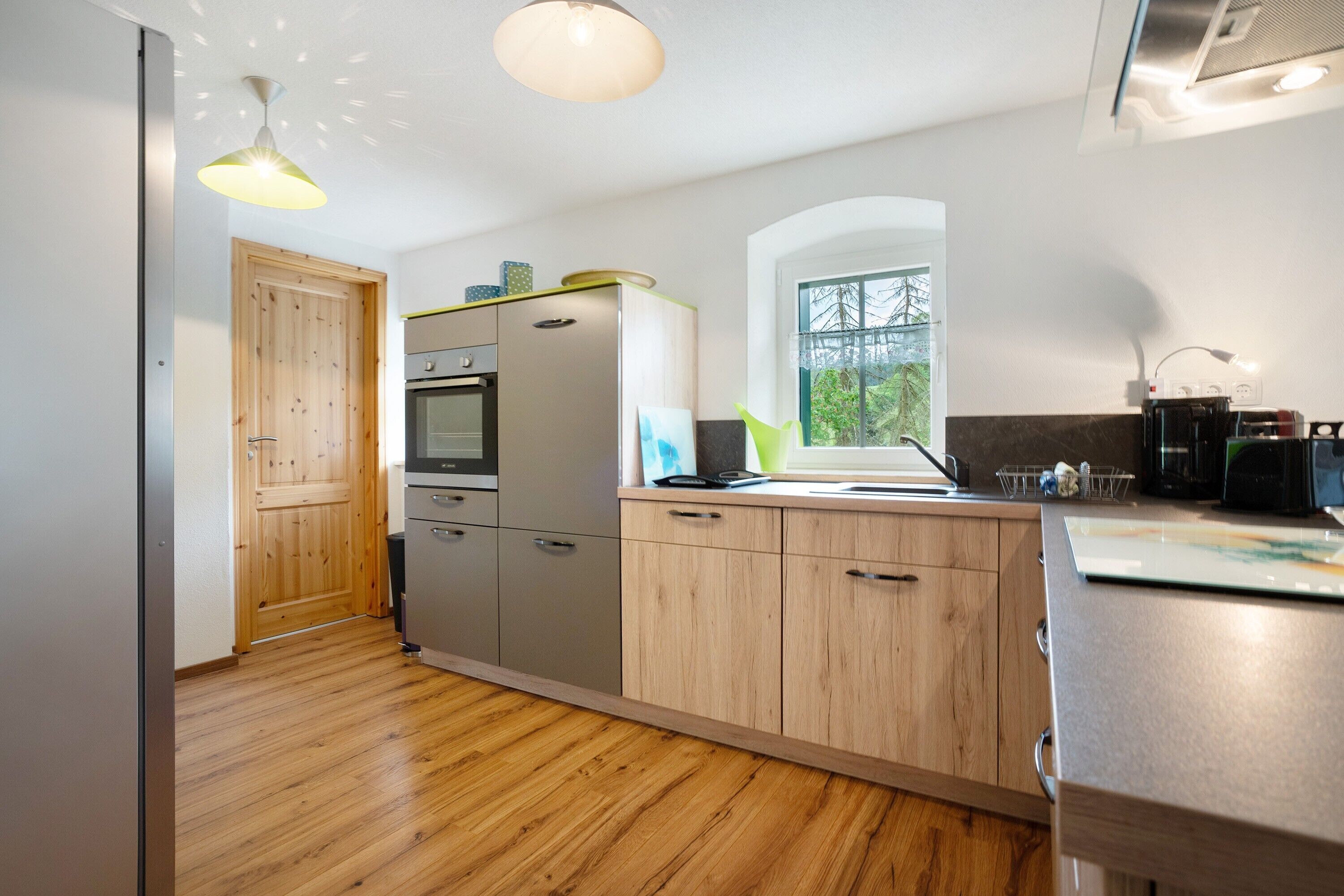 A modern kitchen with wooden cabinets, a stainless steel refrigerator, and a window with a view of trees.