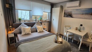 Ein Schlafzimmer mit Bett, Nachttischen, einem Schreibtisch, einem Sessel, einem Fenster mit Jalousien, einer Wand-Klimaanlage und einem Bild mit Strandmotiv.