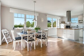 Une cuisine spacieuse avec des meubles blancs, un coin repas avec des chaises blanches et de grandes fenêtres offrant une vue sur la verdure.