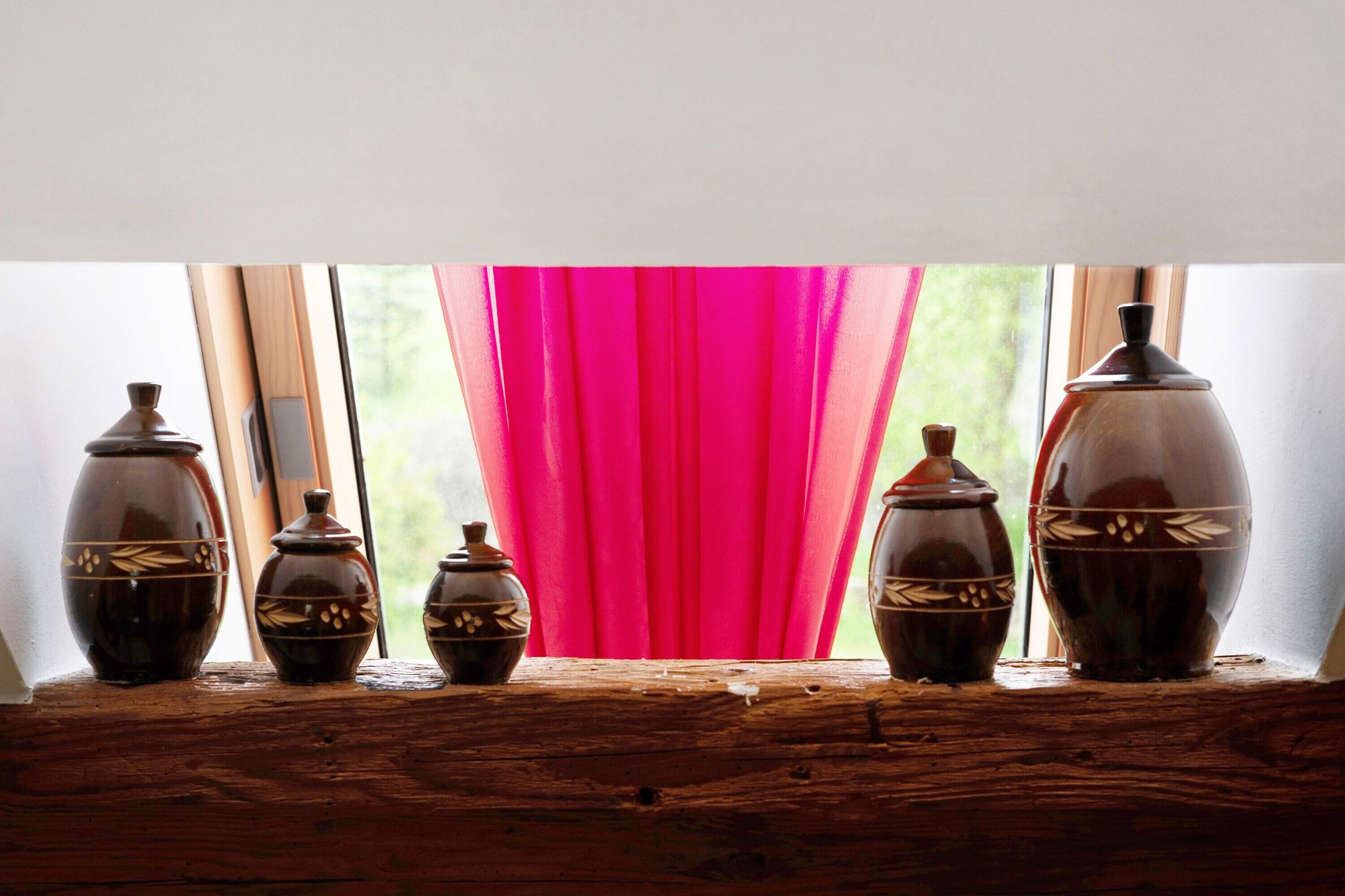 Cinco jarrones de cerámica sobre un repisa de madera junto a una ventana con cortina rosa.