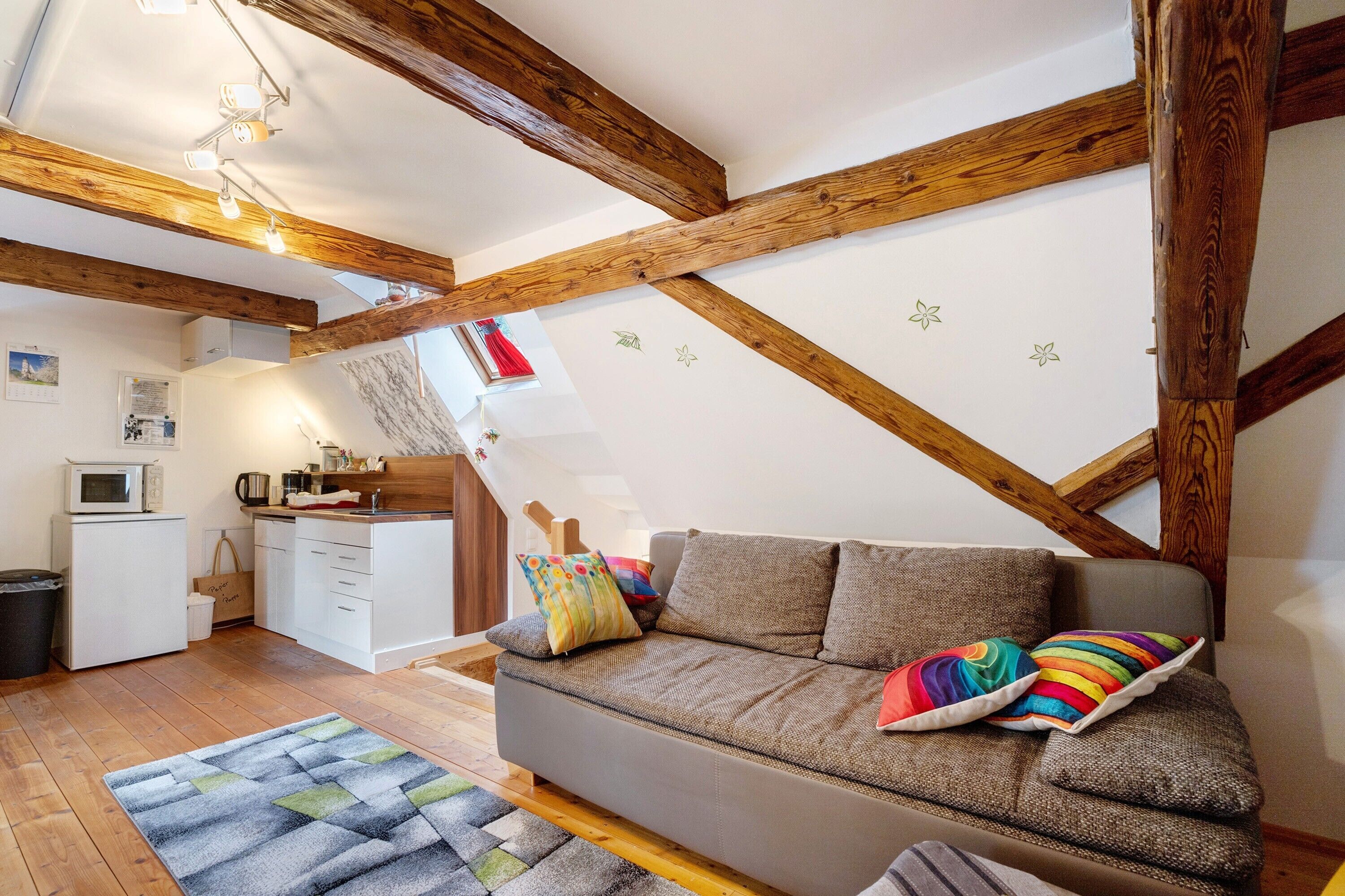A cozy living room with a sofa, wooden beams, a kitchen, and a small white fridge.