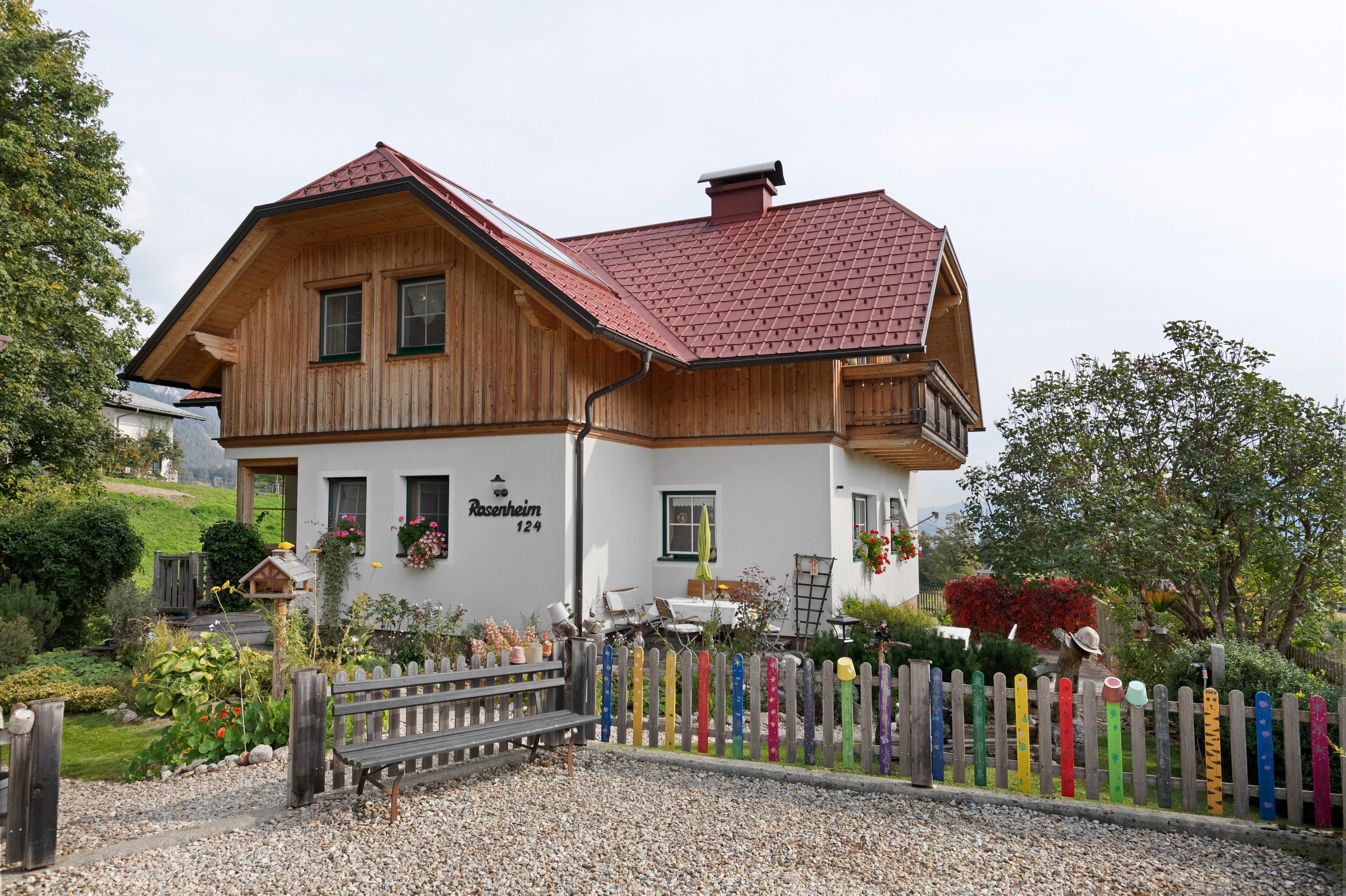 Un bâtiment à deux étages, avec un toit recouvert de tuiles rouges, un revêtement en bois et une façade blanche. On trouve un banc devant le bâtiment, ainsi qu’une clôture colorée avec des poteaux peints.