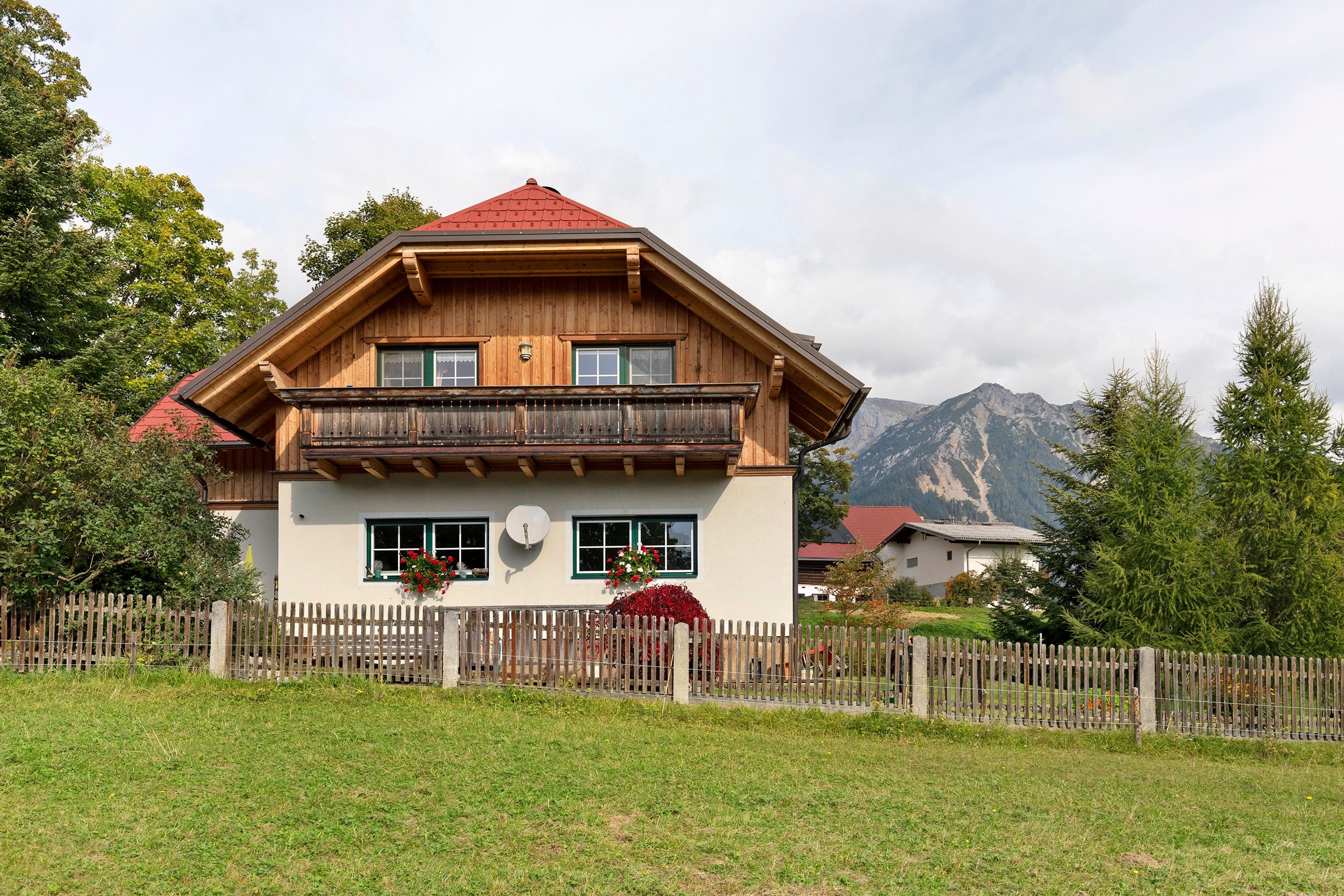 A two-story wooden house with a red roof, a balcony, and a mountain backdrop.