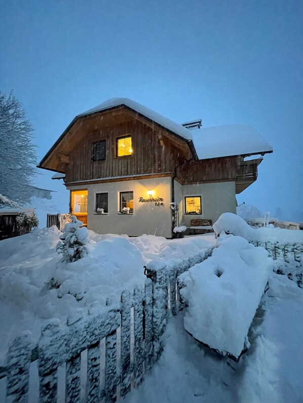 A wooden house with a snow-covered roof and a lit entrance.