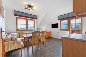 Un salon confortable avec un canapé, une table à manger, des chaises, un téléviseur et de grandes fenêtres donnant sur l’extérieur.