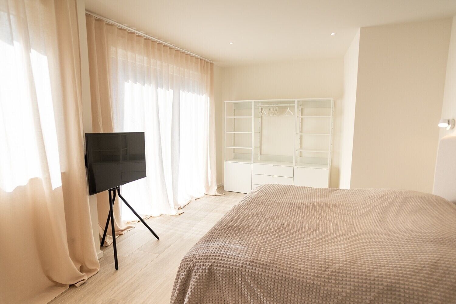 A bedroom with a bed, a TV on a stand, a wardrobe, and a window with curtains.